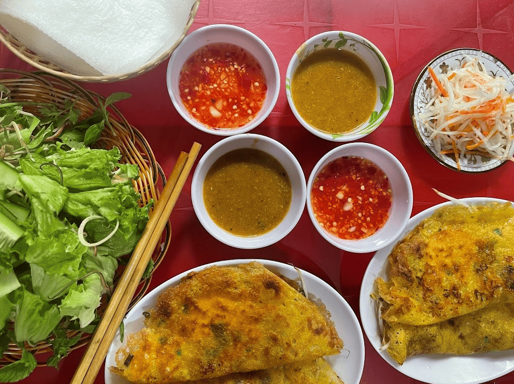  Bàn ăn với bánh xèo vàng giòn, rau sống tươi mát, bánh tráng, dưa góp và nhiều loại nước chấm hấp dẫn