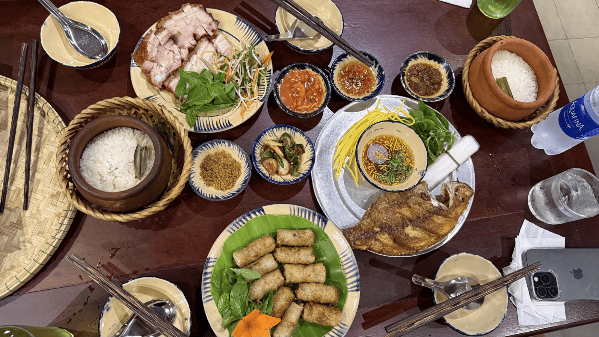Hương vị quê nhà trong mâm cơm truyền thống tại Cơm chạn Huế Bàn ăn gỗ bày biện mâm cơm gia đình với niêu cơm trắng, cá chiên giòn, thịt luộc, nem rán, rau sống và các loại nước chấm, thể hiện sự sum vầy và ấm cúng