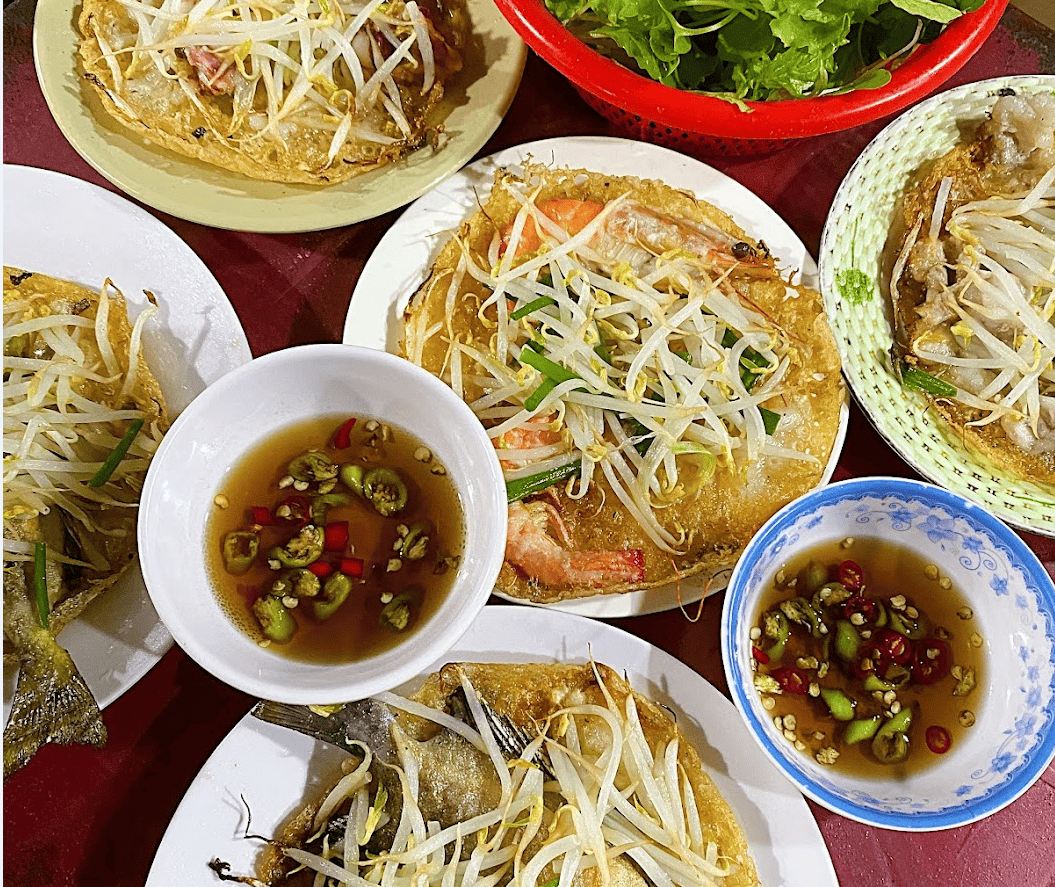 Bàn ăn với nhiều chiếc bánh xèo vàng giòn, nhân tôm thịt tươi ngon, giá đỗ, hành lá, rau sống và nước chấm