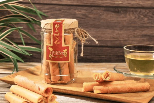 Những chiếc bánh sen chấy cuộn tròn, màu vàng nhạt, giòn rụm, được đựng trong lọ thủy tinh và bày trên khay gỗ, cạnh tách trà thơm.