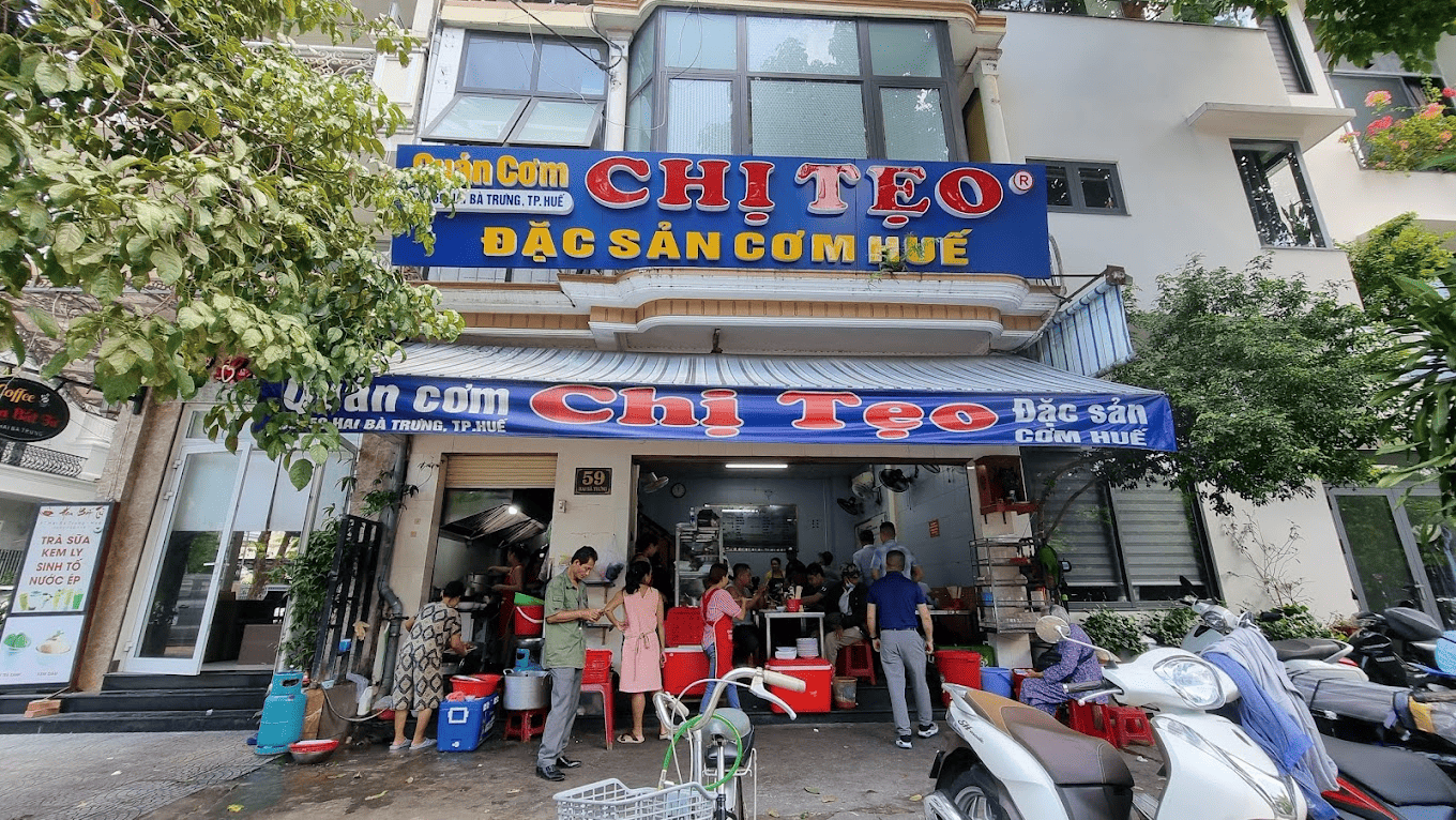 Quán cơm chị Tẹo - điếm đến quen thuộc của người dân địa phương Mặt tiền quán cơm Chị Téo, một địa điểm nổi tiếng với đặc sản cơm Huế tại Hà Nội. Quán có biển hiệu màu xanh nổi bật với tên quán và dòng chữ "Đặc sản cơm Huế"