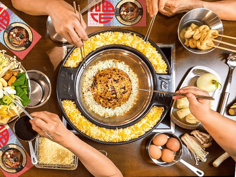 Bàn ăn tại Dookki với nồi lẩu tokbokki phô mai béo ngậy ở giữa, xung quanh là các nguyên liệu tươi ngon và những đôi đũa đang gắp thức ăn đầy hứng khởi.