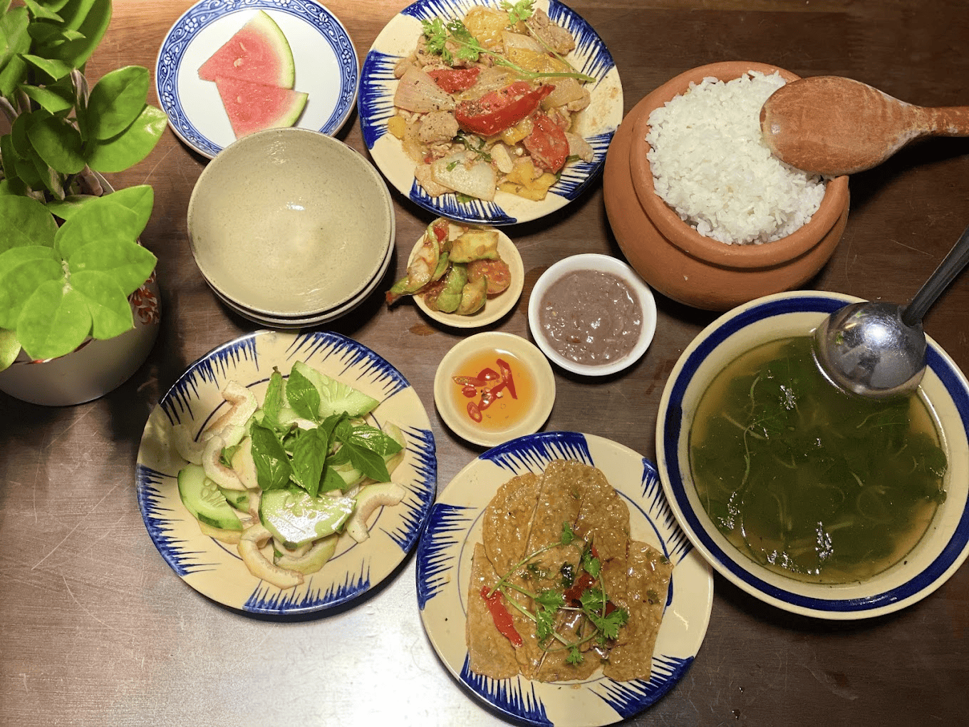 Cà phê Nhà với mâm cơm “chuẩn cơm mẹ nấu Bàn ăn gỗ với niêu cơm trắng dẻo thơm, bát canh rau, đĩa thịt xào, nộm dưa chuột, mắm tôm và dưa hấu, bữa cơm nhà giản dị, đậm đà hương vị quê hương