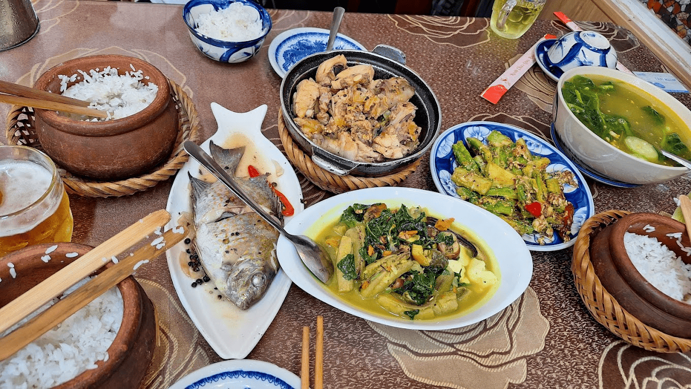 Niêu cơm nóng hổi cùng các món ăn hấp dẫn tại Niêu cơm Lửa Việt Bàn ăn trải khăn họa tiết hoa lá, bày biện mâm cơm gia đình với niêu cơm trắng dẻo thơm, cá diêu hồng chiên, gà kho gừng đậm đà, rau cải xào tỏi xanh mướt