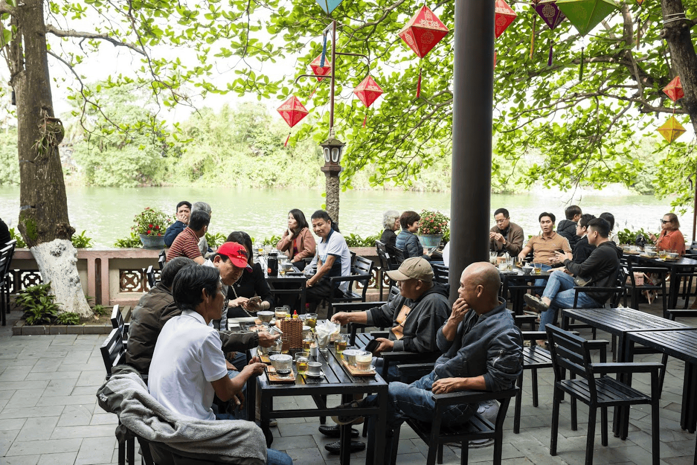 Thực khách dùng bữa trong không gian “rất thơ” tại Nhà hàng Vỹ Dạ Xưa Không gian ngoài trời thoáng đãng của một quán cà phê nằm bên hồ, với nhiều khách đang thưởng thức đồ uống và trò chuyện dưới những chiếc đèn lồng trang trí rực rỡ