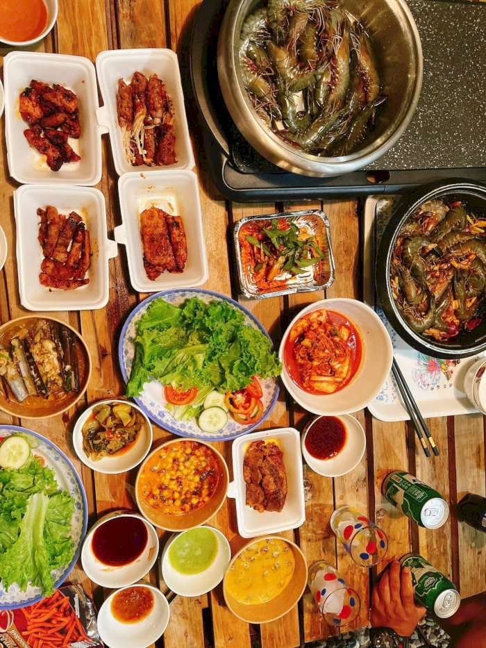 Bàn lẩu có tôm nướng, thịt nướng, rau sống, kim chi và các món ăn kèm hấp dẫn khác, cùng với bia lon và đồ uống.
