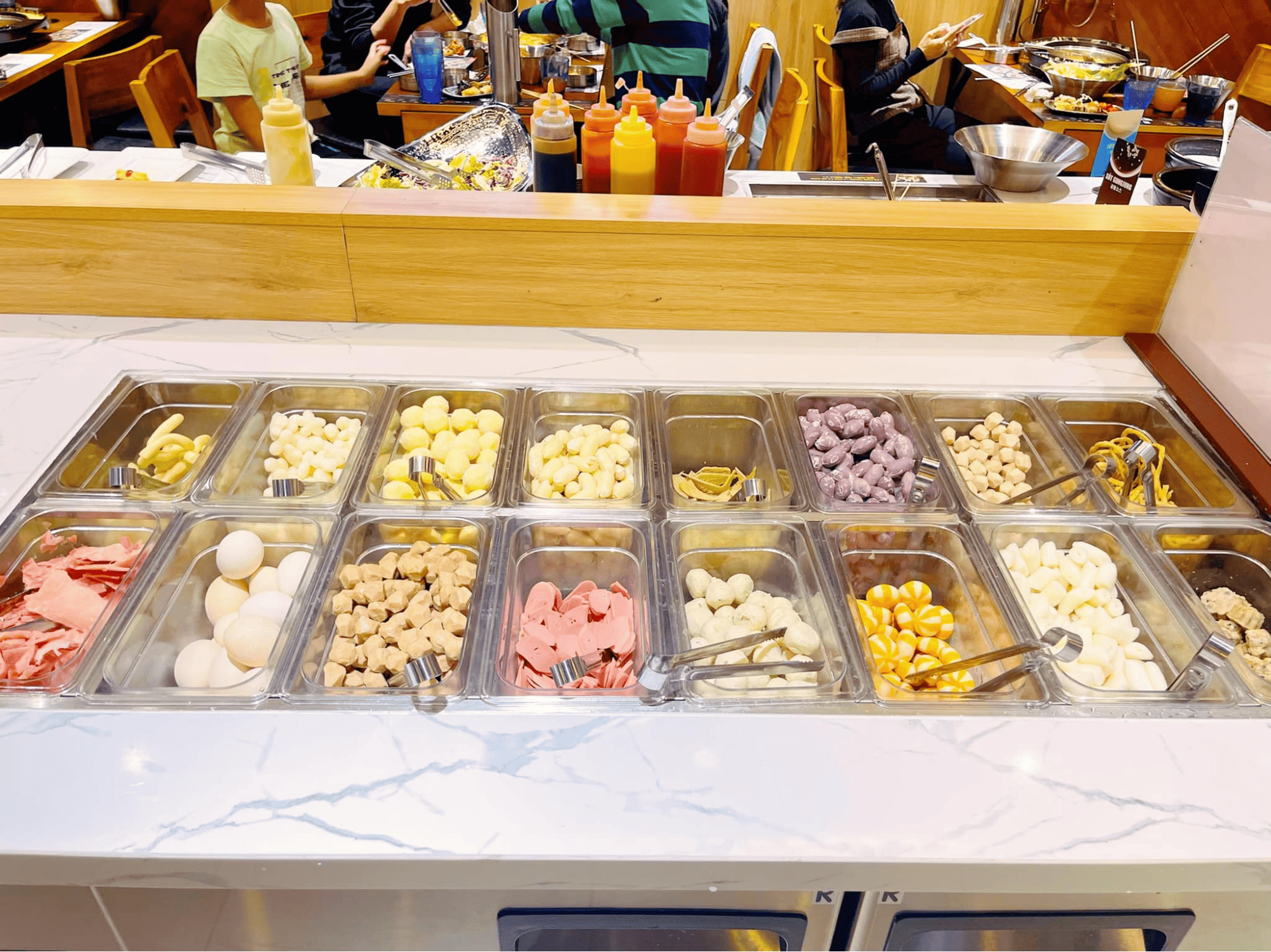 Quầy line tự chọn với vô vàn loại topping hấp dẫn cho món lẩu tokbokki, bao gồm chả cá, xúc xích, bánh gạo, nấm và nhiều loại viên thả lẩu đa dạng màu sắc.